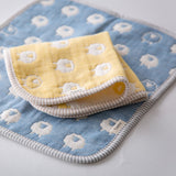 A folded, yellow Fuwara multipurpose small muslin on top of a laid-out blue muslin, both made of Japanese cotton gauze. Each one features a pattern of white woven sheep and matching striped trimming.