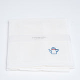 A folded white embroidered muslin cloth with a small blue motif design of a coffee pot in the corner. Made by Harada Orimono and available in NiMi Projects UK.