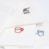 Closeup of three white, embroidered motif muslin cloths, one with a brown coffee mill motif, another with a red coffee cup and the last with a blue coffee pot. Made in Japan by Harada Orimono and available at NiMi Projects UK.