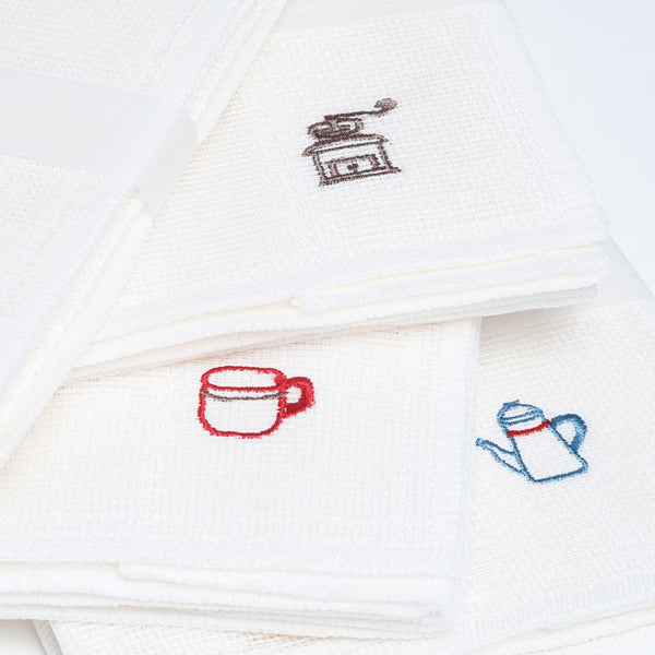 Closeup of three white, embroidered motif muslin cloths, one with a brown coffee mill motif, another with a red coffee cup and the last with a blue coffee pot. Made in Japan by Harada Orimono and available at NiMi Projects UK.