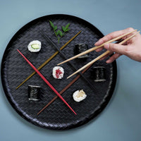 Sushi arranged on a black woven tray with crossed Style of Japan double-end ryokuchi hashi chopsticks, with a hand holding chopsticks above.