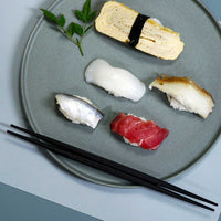 A pair of dark brown hashi chopsticks that are tapered at both ends placed on the edge or a round, grey-blue plate with various pieces of sushi.