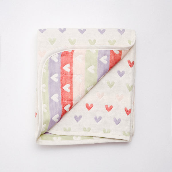 A folded, reversible Japanese off-white cotton muslin blanket patterned with rows of multicolour hearts. One corner is folded upward to show both sides of the fabric. Available at NiMi Projects UK and made by Fuwara in Japan.	