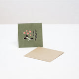 A square Japanese Motif Mini Card, featuring an illustration of shochikubai, a Japanese motif of plum blosson, bamboo and pine on a green background. Displayed with a brown envelope. Printed in Japan and available at NiMi Projects UK.