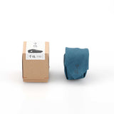 The Kuon Madoka acorn-shaped singing bowl chime cardboard box packaging, featuring the Kisen brand logo, displayed next to a chime wrapped in a blue cotton cleaning cloth. Designed by Yotsukawa Seisakusho for its Kisen brand.