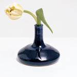 A Maki Baxter pod vase in blue glaze, available at NiMi Projects, featuring a round flat bottom and tall thin neck, and holding a single yellow tulip.