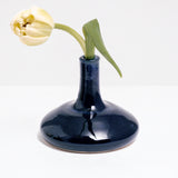 A Maki Baxter pod vase in blue glaze, available at NiMi Projects, featuring a round flat bottom and tall thin neck, and holding a single yellow tulip.