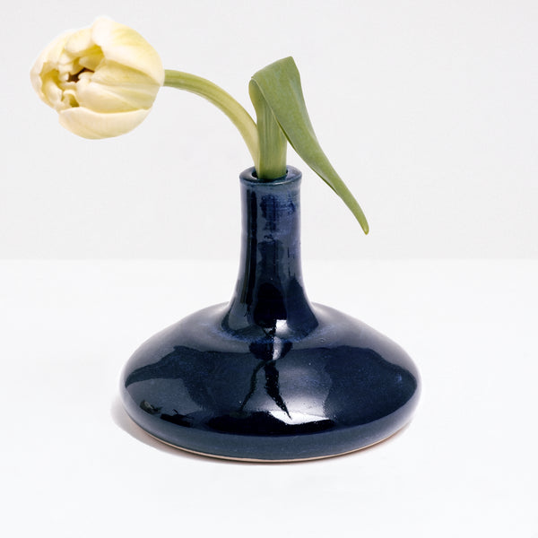 A Maki Baxter pod vase in blue glaze, available at NiMi Projects, featuring a round flat bottom and tall thin neck, and holding a single yellow tulip.