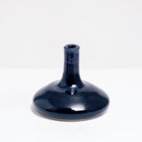 A small blue-glazed, flat, round-bottomed vase, with a tall slim neck, designed and made by Maki Baxter and available at NiMi Projects UK.