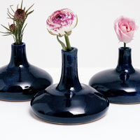 Three blue-glazed mini pod vases, designed and handmade by ceramicist Maki Baxter, each featuring flattened round bottoms and tall slim necks and holding single stemmed pink flowers. Available at NiMi Projects UK.