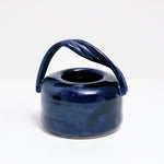 A blue pot-shaped ceramic vase, designed and made by Maki Baxter, featuring a twisted handle on show at NiMi Projects UK.