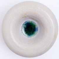 A top view of a large, rotund, donut-shaped ceramic decorative dish in grey with a pool of green-blue crackled glaze at the bottom, hand made by Maki Baxter and on show at NiMi Projects UK.
