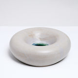 A side-view of a large, rotund, donut-shaped ceramic decorative dish in grey with a pool of green-blue crackled glaze at the bottom, hand made by Maki Baxter and on show at NiMi Projects UK.