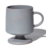 A soft grey, lightly speckeled, satin-glazed stemmed (or footed) mug with a round base and curved handle. Hand made in Japan by Angle and available at NiMi Projects UK.
