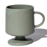 A sage green, satin-glazed stemmed (or footed) mug with a round base and curved handle. Hand made in Japan by Angle and available at NiMi Projects UK.