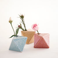 Three diamond-shaped, Japanese ceramic bud vases in salmon pink, yellow and blue, each holding a small flower. Available at NiMi Projects UK.