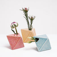 Three diamond-shaped, Japanese ceramic bud vases in salmon pink, yellow and blue, each holding a small flower. Available at NiMi  Projects UK. 
