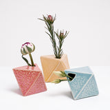 Three diamond-shaped, Japanese ceramic bud vases in salmon pink, yellow and blue, each holding a small flower. Available at NiMi  Projects UK. 