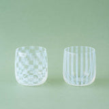 Two clear, Japanese glass cups, made by Hirota Glass, displayed against a mint green background. Each glass features translucent white aburidashi geometric patterns — one vertical stripes, the other checked — and is designed by Moheim.