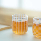 Two clear, Japanese glass cups, made by Hirota Glass, filled with ice tea and displayed on a white tray. Each glass features translucent white aburidashi geometric patterns — one vertical stripes, the other checked — and is designed by Moheim.