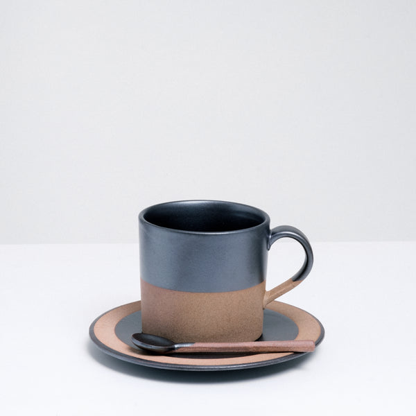 An earthenware Angle Soil mug, made in Japan and dipped in a satin black glaze. Pictured at NiMi Projects UK with a matching earthenware spoon, both placed on top of a matching Angle Soil saucer.