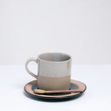 An earthenware Angle Soil mug, made in Japan and dipped in a glossy grey glaze. Pictured at NiMi Projects UK with a matching earthenware spoon, both placed on top of a matching Angle Soil saucer.