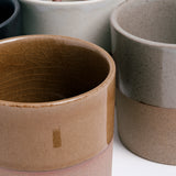 Closeup detail of several Angle ceramic Soil mugs showcasing their dipped glazes in taupe, grey and white and their lower halvesof exposed orange earthenware clay. Available at NiMi Projects UK.