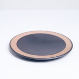 A  mino-yaki earthenware small plate, designed by Angle and made in Japan, featuring a central circle of shimmering graphite black glaze and a matching line of black on its outer rim. An exposed, unglazed clay border of the plate showcases the speckled texture of the raw orange clay. Available at NiMi Projects UK.