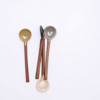 A group of four Angle ceramics mino-yaki pottery tea spoons on display at NiMi Projects UK. Each spoon has an exposed orange clay handle and a head dipped in a rustic coloured glaze. From left to right: a glossy yellow version, graphite black, satin white and glossy grey.