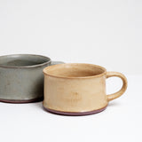 Two low and wide, flat ceramic mugs; one with a green gray country glaze and the other in a beige yellow. Made in Japan by Angle and available at NiMi Projects UK.