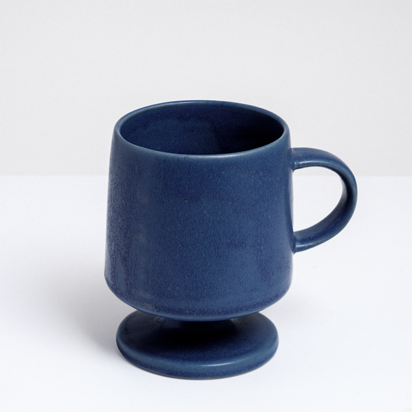 A dusky blue, satin-glazed footed (stemmed) mug with a circular base and curved handle. Hand made in Japan by Angle and available at NiMi Projects UK.