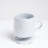 A white, satin-glazed stemmed (or footed) mug with a round base and curved handle. Hand made in Japan by Angle and available at NiMi Projects UK.