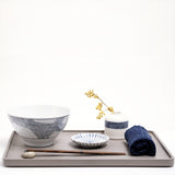 Lifestyle image of an Ito Tsumugi Japanese white porcelain mino-yaki ramen and noodle bowl, featuring a pattern of criss crossed lines, displayed on a grey wooden tray, with a small white dish with blue lines, a bud vase with a patterned band of blue lines and a rolled blue Japanese gauze napkin. On display at NiMi Projects UK.
