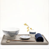 Lifestyle image of an Ito Tsumugi Japanese white porcelain mino-yaki ramen and noodle bowl, featuring a pattern of criss crossed lines, displayed on a grey wooden tray, with a small white dish with blue lines, a bud vase with a patterned band of blue lines and a rolled blue Japanese gauze napkin. On display at NiMi Projects UK.