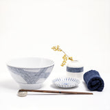 Lifestyle image of an Ito Tsumugi Japanese white porcelain mino-yaki ramen and noodle bowl, featuring a pattern of criss crossed lines, displayed with a small white dish with blue lines, a bud vase with a patterned band of blue lines and a rolled blue Japanese gauze napkin. On display at NiMi Projects UK.