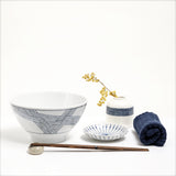 Lifestyle image of an Ito Tsumugi Japanese white porcelain mino-yaki ramen and noodle bowl, featuring a pattern of criss crossed lines, displayed with a small white dish with blue lines, a bud vase with a patterned band of blue lines and a rolled blue Japanese gauze napkin. On display at NiMi Projects UK.