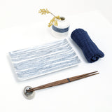 The Angle Ito Tsumugi rectangular sushi plate, showing its pattern of fine blue lines. Displayed with a pair of wooden hashi chopsticks, a vase with a blue band of striped patterning and a rolled Japanese gauze napkin. Made in Japan by Angle and available at NiMi Projects UK.