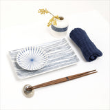 An Angle Japanese porcelain Ito Tsumugi rectangular sushi plate, featuring a pattern of blue fine lines, made in Japan and displayed with a pair of wooden hashi chopsticks, a white bud vase with patterned band of blue lines, and a rolled Japanese gauze napkin. Available at NiMi Projects UK.