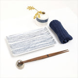 An Ito Tsumugi rectangular sushi plate, featuring a pattern of blue fine lines, made by Angle in Japan and displayed with a pair of wooden hashi chopsticks, a white bud vase with patterned band of blue lines, and a rolled Japanese gauze napkin. On show at NiMi Projects UK.