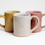 Three glazed ceramic mugs — yellow, white and pink — each with their handles positioned at an angle, designed to make it easier to hold and tilt. Hand made by Angle in Japan and available at NiMi Projects UK