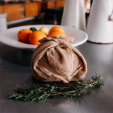 A small bundle wrapped in a Ban Inoue organic cotton kitchen cloth, placed on a dark tabletop, with a plate of citrus fruits and kitchen items blurred in the background.
