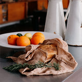 A rustic loaf of bread wrapped in Ban Inoue, clove brown, organic cotton muslin cloth, styled on a dark countertop with rosemary sprigs, a white plate of oranges, and two white enamel pitchers in the background.