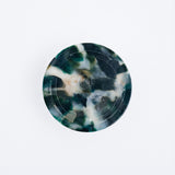 A recycled marine plastic coaster made by Buoy in Japan and available at NiMi Projects. The coaster's mottled black colour includes flecks of white and deep green, and is produced by the original colours of plastics waste collected from the coasts of Japan.