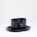 Two black semi-circle cups, made by Ceramic Japan, that sit together with their flat sides meeting on a shared double saucer, on show at NiMi Projects UK.