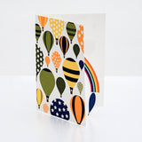 A Chisato Tamabayashi hot air balloon 3d popup greeting card, showing its front graphic illustration of various balloons in orange, yellow, green and blue with patterns of strips and stars. The card is slightly opened to reveal a popup rainbow with a blue balloon hanging from it's end. On show at NiMi Projects UK. 