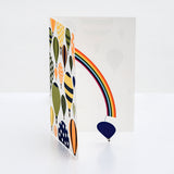 A Chisato Tamabayashi hot air balloon 3D popup greeting card, showing a graphic illustration of various balloons in orange, yellow, green and blue with patterns of strips and stars. The card is opened to reveal a popup rainbow with a blue balloon hanging on a thread from it's end. Available at NiMi Projects UK. 