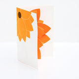 A Chisato Tamabayashi orange flower 3D popup greeting card, showing a partial graphic of bright orange flower on the top left corner on a white background, and slightly opened to reveal the petals of the orange popup flower inside. On show at NiMi Projects UK. 