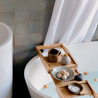 A bamboo bath tray resting on a white designer bath with a backdrop of a grey tiled wall. On the tray is a stoneware mug, a grey bud vase with a yellow flower, a duck-egg blue bowl with five hinoki (Japanese cypress) bath balls, a rock and a small bowl of bath salt. Photo taken by NiMi Projects UK.