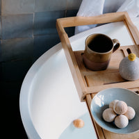 A closeup shot of a bamboo bath tray resting on a white designer bath with a backdrop of a grey tiled wall. On the tray is a stoneware mug, a grey bud vase with a yellow flower and a duck-egg blue bowl with five hinoki (Japanese cypress) bath balls. Photo taken by NiMi Projects UK.