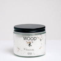 A short, cylindrical glass jar of bath salts with a flat, black, metal screwtop lid. The jar has WOOD written on it, and the Japanese kanji for wood, with "homework" written below. Available at NiMi Projects UK.